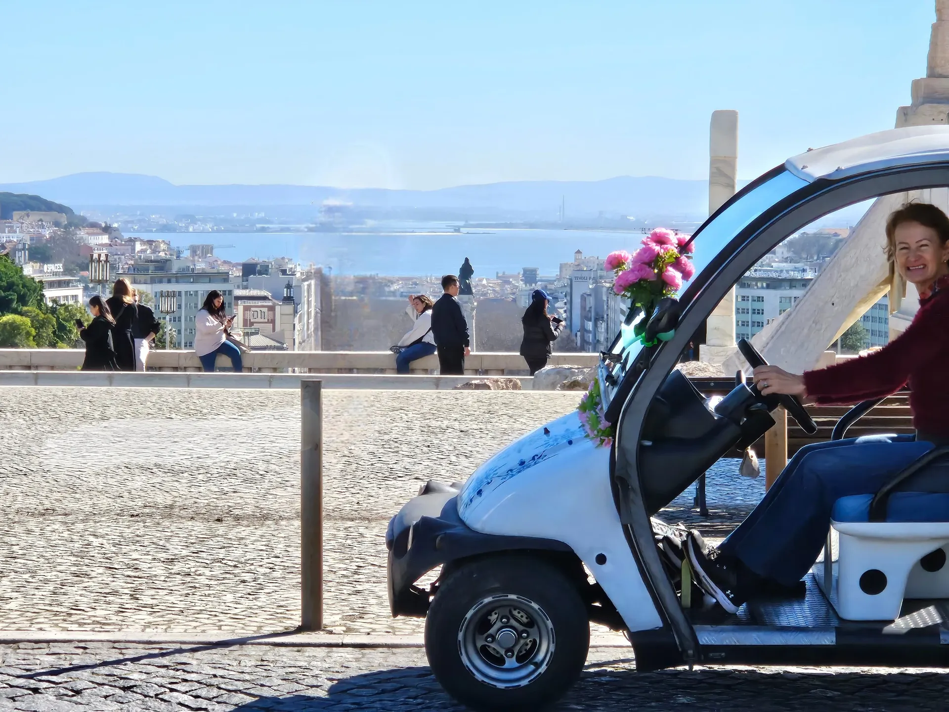 Tuk-tuk azul e branco decorado com flores e a guia Susane sorridente com a deslumbrante paisagem de Lisboa ao fundo